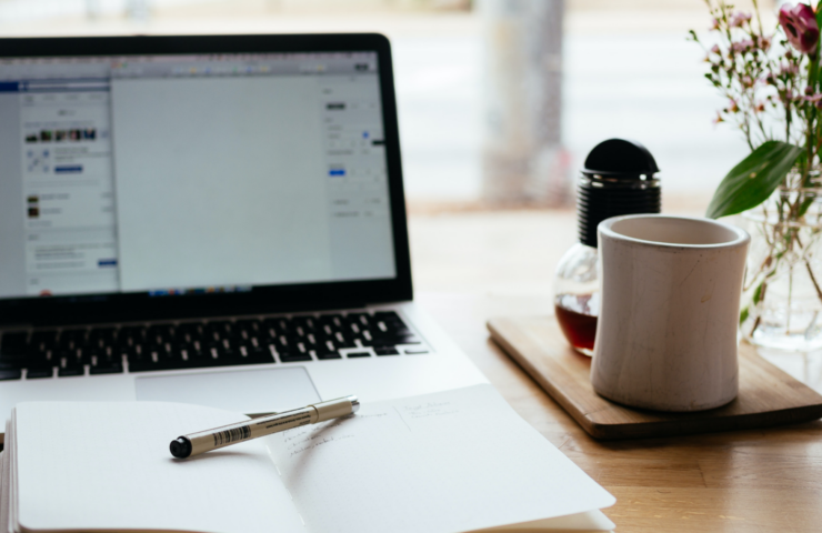 An image of a coffee mug and a notebook on a desk with a laptop